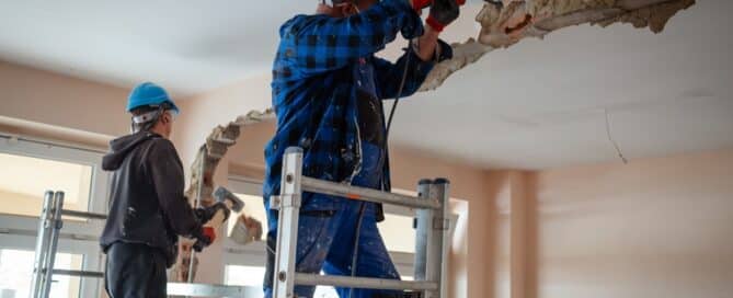 Two Construction Workers On Scaffolding Demolishes A Wall
